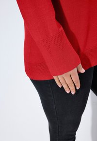 Maglione rosso a maglia con orlo e maniche strutturati, caratterizzato da ritagli triangolari. Indossato sopra pantaloni neri aderenti. Tessuto liscio con una finitura morbida.