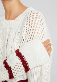 Weißer gestrickter Pullover mit offenem Netzmuster, ausgestattet mit roten horizontalen Streifen an den Ärmeln, gerippten Bündchen und einem strukturierten Halsausschnitt.