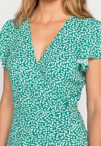 Grünes Blumen-Wickelkleid mit V-Ausschnitt, kurzen, fließenden Ärmeln und einem Bindegürtel in der Taille; der Stoff hat eine leichte Textur.