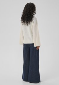 Pull-over taille XL de couleur crème avec des poignets côtelés, associé à un jean en denim foncé à jambes larges. Coupe décontractée et texture douce visibles.