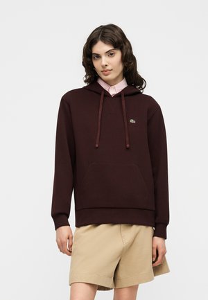 Junge Person mit welligem dunklem Haar trägt einen dunkelbraunen Hoodie über einem pinken Hemd mit Kragen und beigen Shorts und steht vor einem schlichten Hintergrund.