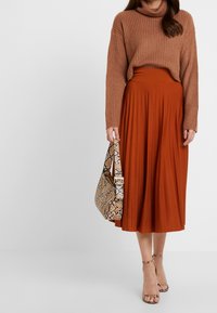 Pull à col roulé marron côtelé, jupe midi plissée couleur rouille, sac à main imprimé serpent, et sandales à talons avec lanières. Accent sur les tons terreux et les textures.