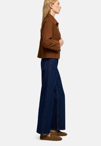 Brązowa zamszowa kurtka, ciemnoniebieskie szerokie dżinsy oraz brązowe sznurowane buty. Model zwrócony w lewo, prezentujący casualowy strój.