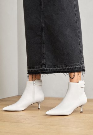 Bottines blanches à bout pointu avec talons kitten portées avec un pantalon large à ourlet effiloché noir sur un sol en bois clair.
