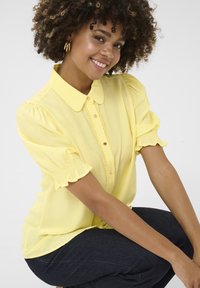 Blusa gialla con scollatura a collare, maniche a palloncino e dettaglio dei bottoni. Realizzata in tessuto morbido con dettagli plissettati e vestibilità comoda.