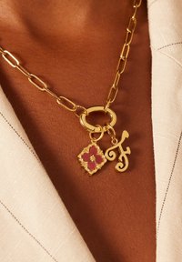 Gouden ketting met een ronde schakel en twee bedels: een bloemenontwerp met een roze centrum en een gestileerde letter "F" bedel.