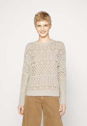 Pull en tricot beige clair présentant un motif de points ouverts, un col rond et des manches longues, associé à un pantalon ample de couleur beige.