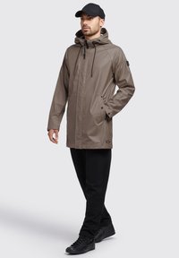 Vandtæt khaki-jakke med hætte, der har sorte snore og sidelommer, båret med sorte bukser og sneakers. Glat tekstur.