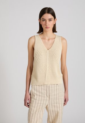 Giovane donna che indossa un top in maglia crema senza maniche e pantaloni beige chiaro con sottili righe verticali nere, in piedi davanti a uno sfondo chiaro uniforme.