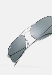Sunglasses with a silver metal frame, dark grey lenses, and adjustable nose pads; features angular shape and minimalistic design.