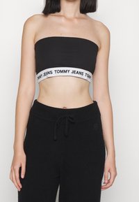 Czarny top bez ramiączek o kroju crop z białą elastyczną taśmą z logo "TOMMY JEANS". Tkanina wydaje się miękka i gładka, z dopasowanym krojem.