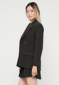 Jong vrouw met lang ombre haar in een donker gemuste blazer en een geplooide zwarte rok, staande zijwaarts tegen een witte achtergrond.