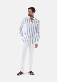 Camicia di lino leggera con strisce verticali azzurre e bianche, davanti con bottoni, abbinata a pantaloni bianchi e sandali scuri.