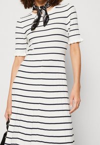 Frau, die ein weißes Kleid mit horizontalen schwarzen Streifen trägt, kurze Ärmel mit gerafften Rändern, eine Perlenkette und einen schwarzen gemusterten Halstuch.