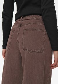 Rjave denim hlače z visokim pasom in ohlapnim krojem, z dvema zadnjima žepoma in kontrastnim šivanjem. Tkanina ima gladko, teksturirano površino.