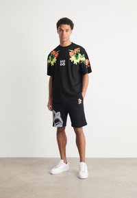 T-shirt noire surdimensionnée avec impression florale colorée sur les épaules et logo "HUGO". Short noir avec un graphique de raton laveur et détail de logo. Baskets blanches.
