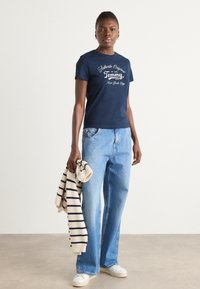 Mørkeblå bomulls t-skjorte med hvit trykt tekst, kombinert med lyseblå vide jeans og en kremfarget stripete genser over armen.
