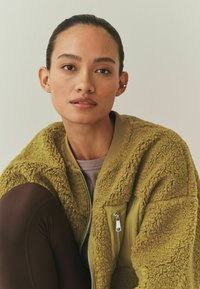 Mlada ženska z zategnjenimi lasmi, oblečena v zeleno teksturirano flis jakno in temne legice, sedi ob enostavni svetli podlagi.