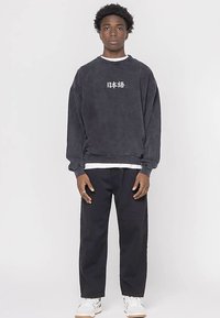 Tamnosiva majica dugih rukava s bijelim japanskim tekstom, okruglim izrezom, širokog kroja i rebrastim manžetnama, uparena s crnim hlačama i bijelim tenisicama.