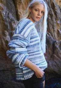 Mladá žena s dlhými blond vlasmi oblečená v modro-bielom pruhovanom pletenom svetri stojaca vonku na pozadí skalnatého terénu.