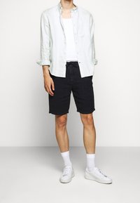 Ljusgrön linneskjorta med knappar över en vit linne, parat med mörka shorts och vita sneakers, med vita strumpor synliga.