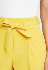 Gele broeken met een stoffen riem die in een strik om de taille is geknoopt. Gladde textuur en wijde pijpen met plooien aan de voorkant voor extra detail.