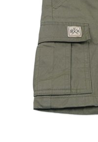 Vihreät kangascargo-housut, joissa on tikkupussi beige etiketillä. Teksturoitu materiaali ja ompeleita saumoissa.