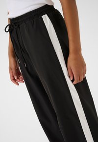 Zwarte sportbroek met een witte zijstreep, voorzien van een elastische tailleband en een verstelbaar trekkoord. Glad stoffen textuur zichtbaar.