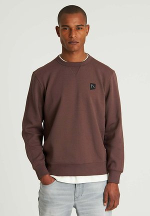Brun sweatshirt med ribbade muddar och fåll, med en liten svart logotyp på bröstet. Bärs över en vit skjorta och kombineras med ljusa jeans.