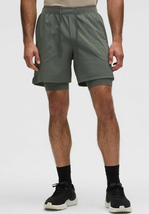 Mand iført olivengrønne lagdelte træningsshorts, sorte sokker og sorte sneakers, stående mod en ensfarvet baggrund.