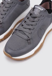 Zapatillas de malla gris transpirables con acentos sintéticos, cuello acolchado y cordones planos. Media suela blanca y suela externa de goma marrón texturizada.