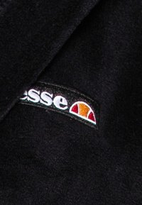 Juodas fliso audinys su siuvinėtu logotipu, kuriame yra baltas tekstas ir spalvota pusapvalė forma raudonos, oranžinės ir geltonos spalvos akcentais.