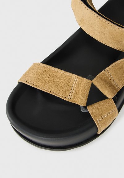 Černé sandály s hnědými semišovými pásky, které mají nastavitelné uzávěry na suchý zip přes prsty a kolem kotníků.