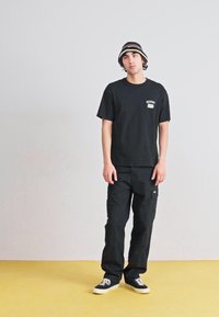 T-shirt en coton noir avec un logo sur la poitrine, associé à un pantalon cargo noir avec des poches latérales. Porté avec un chapeau seau noir et blanc.