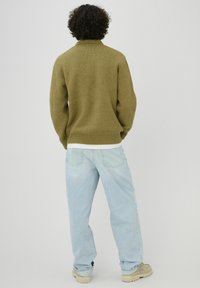 Personne aux cheveux bouclés de dos, portant un pull vert olive, un jean bleu clair et des chaussures beiges, sur un fond uni.