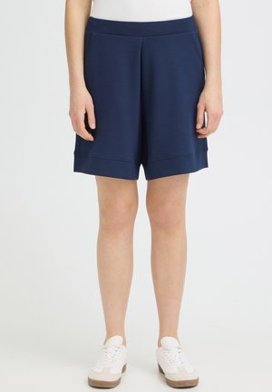 Persoană care poartă pantaloni scurți bleumarin până la genunchi, adidași albi și un top alb, stând în fața unui fundal alb simplu.