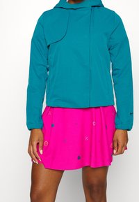 En teal jacka med huva och en fickdetalj, som används tillsammans med en rosa kjol med ett mönster av små ikoner och en utsvängd design.