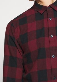 Chemise à carreaux rouge et noir avec un col, présentant un tissu texturé et des boutons noirs le long de l'avant.