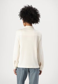 Kremfarget langermet skjorte laget av mykt stoff, med krage, knappede mansjetter og sidesplitter, kombinert med lyseblå jeans.