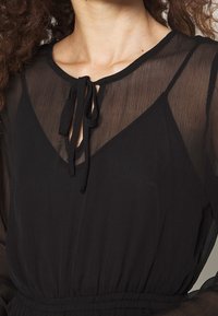 Nära bild av en kvinna som bär en svart genomskinlig blus med en knuten halsringning och en svart linne under.