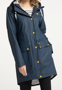 Mörkblå regnjacka med huva, prydd med gula knappar, framfickor och ett randigt innerfoder, tillverkad av vattentätt material.