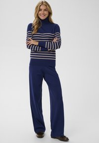 Marineblauer Rollkragenpullover mit beigen horizontalen Streifen, kombiniert mit lockeren marineblauen Hosen. Beide Kleidungsstücke haben eine glatte Textur und eine entspannte Passform.