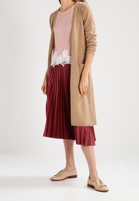 Femme portant un long cardigan beige, un haut rose avec un ourlet en dentelle blanche, une jupe midi plissée bordeaux et des mocassins beiges à pompons, debout sur un sol blanc.