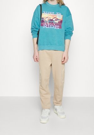 Person trägt einen türkisfarbenen Pullover mit der Aufschrift "WILD RIDE" und Pferde-Grafik, beige Hose, weiße Sneakers und eine schwarze Umhängetasche.
