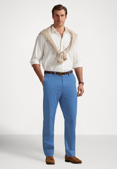Roupa masculina composta por uma camisa branca com botões, calças azuis claras, suéter bege pendurado ao redor do pescoço e sapatos sociais castanhos.