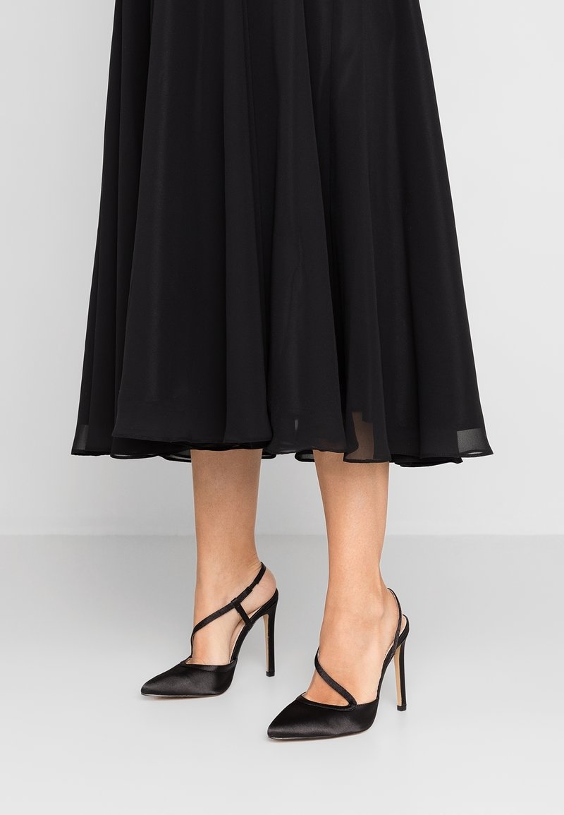 Chaussures à talons hauts en satin noir avec bout pointu, dotées d'une bride à la cheville et de détails délicats à l'avant, associées à une robe noire fluide.