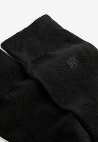 Schwarze Socken aus weichem Material, mit einem gerippten Bündchen und einem kleinen, in derselben Farbe gestickten Logo. Glatte Textur überall.