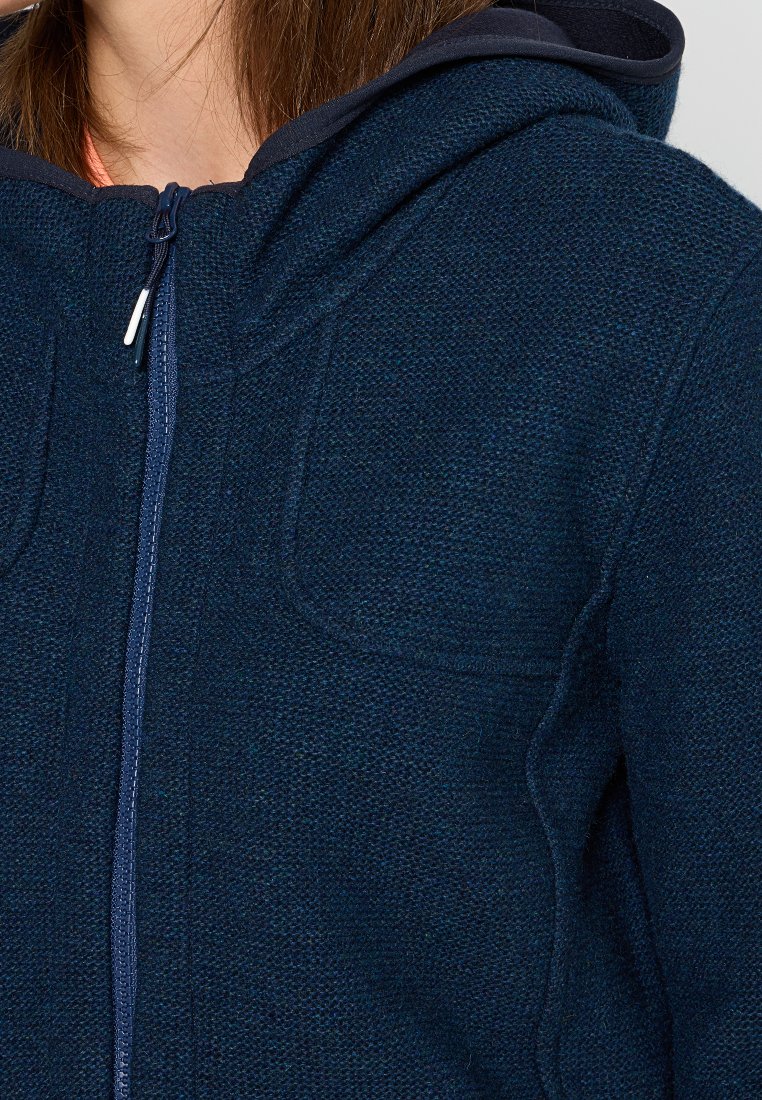 Ravvicinato di una persona che indossa una felpa con zip di colore blu navy, con una texture visibile e un cappuccio, e una maglietta parziale di colore corallo sotto.