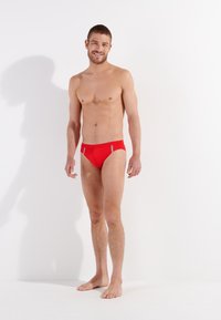 Červené plavky ve fitted designu, s kontrastními bočními panely. Tkanina vypadá hladce, s minimalistickým a sportovním vzhledem.