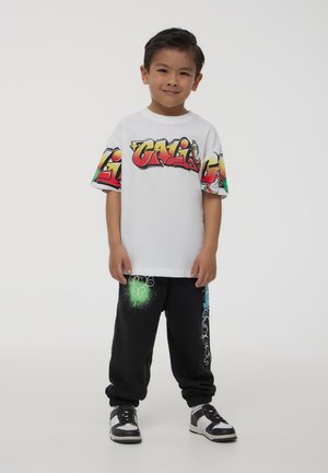 T-shirt bianca con testo in stile graffiti colorato; pantaloni sportivi neri con accenti grafici verdi e blu; sneakers in bianco e nero.